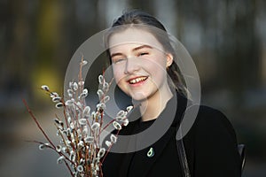 Close up emotional portrait of young happy beautiful woman with a bouquet of pussy-willows wearing black coat strolling at evening