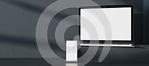Close up of empty white laptop and cellphone on dark gray desk. Concrete wall background, shadows and pedestal or podium. Device