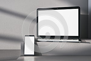 Close up of empty white laptop computer and phone on gray desk. Concrete wall background, shadows and pedestal or podium. Device