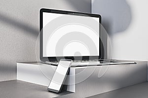 Close up of empty white laptop and smartphone on gray desk. Concrete wall background, shadows and pedestal or podium. Device