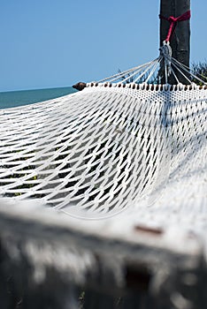 Close up of an empty white rope hammock hanging with trees in a shadow of tree branches on a tropical beach at noon.