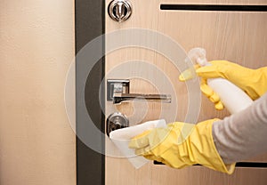 Close up entrance door knob being cleaned with sanitizer spray by female hands in yellow latex gloves, selective focus