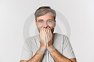 Close-up of excited and surprised man, cover mouth and giggle from amazement, standing over white background