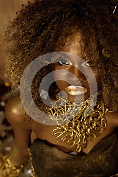 Close up of Exotic African American woman with curly afro hairstyle wearing dark makeup and gold accessories looking directly at