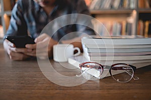 Close up eyeglasses and books stack on wooden desk in university or public library with man using mobile phone reading or text