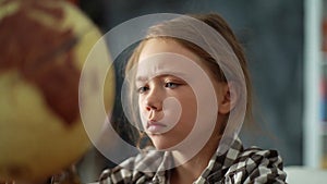 Close-up face of adorable thoughtful child girl spinning small globe with interest, looking at countries of world.