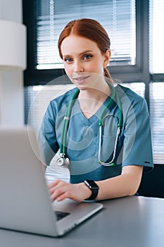 Close-up face of attractive smiling young female doctor in blue green medical uniform typing on laptop computer, sitting