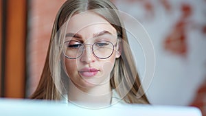 Close-up face of beautiful busy young business woman wearing glasses looking at screen of laptop