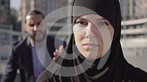 Close-up face of beautiful muslim woman with sore eyes looking at camera as blurred man gesturing and yelling at the