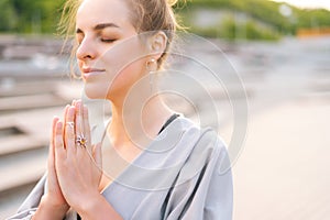 Close-up face of calm attractive young woman practicing yoga performing namaste pose with closed eyes outside in city