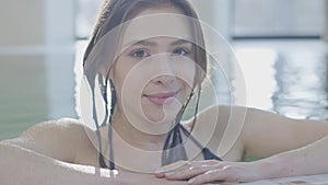 Close-up face of charming brunette woman posing at poolside and smiling at camera. Portrait of beautiful positive girl