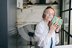 Close-up face of cheerful redhead young woman gently cradling surprise gift box to face at home.