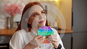 Close-up face of dreaming relaxed redhead young woman pushing colorful iridescent soft silicone bubbles sitting at table