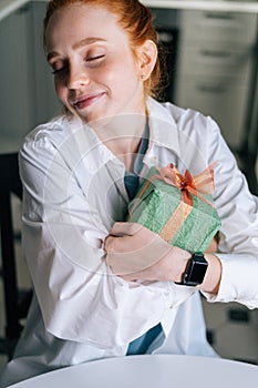 Close-up face of dreamy redhead young woman gently cradling surprise gift box to heart at home.