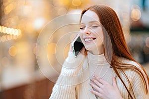 Close-up face of excited smiling young woman talking on mobile phone with closed eyes standing in shopping mall with