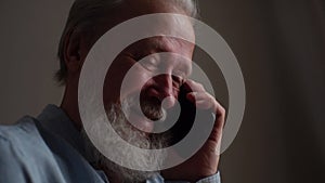 Close-up face of happy gray-haired senior adult male with tears in eyes talking on mobile phone with family at home.