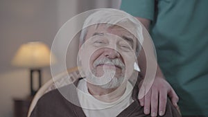 Close-up face of happy old man looking at camera. Unrecognizable male nurse holding hand on man`s shoulder. Portrait of