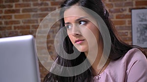 Close-up face of intelligent pretty caucasian female with brown eyes and thoughtful look, using her laptop while sitting