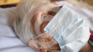 Close up face of old woman wearing protective mask from virus. Sick lady with sad sight and medical face mask lying at