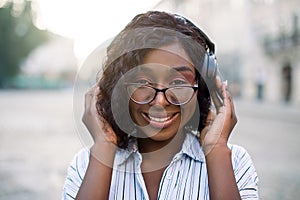 Close up face portrait of pretty smiling young african lady, listening to music on headphones, enjoying walk outdoors on