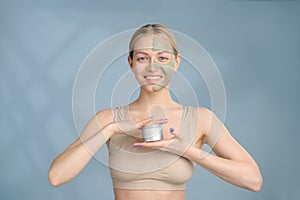 A close-up face portrait of a young blonde woman. Green beauty mask on half of the face. A woman holds a jar of