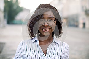 Close up face portrait of young smiling pretty African woman wearing eyeglasses and striped shirt, posing to camera