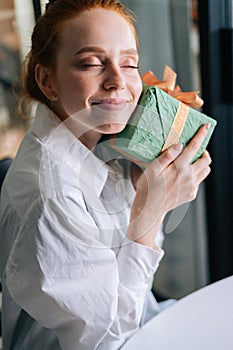 Close-up face of pretty redhead young woman gently cradling surprise gift box to face at home.