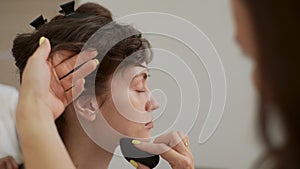 Close-up face of pretty young woman being given makeup and hairstyle in beauty salon. Makeup artist applying fluid