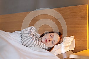 Close-up face of pretty young woman sleeping well in bed hugging soft white pillow at home, bedside lamp lighting with