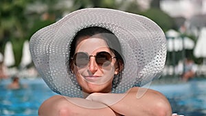 Close-up face relaxed smiling woman in sunglasses and white hat enjoying sunbathing at swimming pool