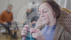 Close-up face of serious elderly Caucasian woman knitting in armchair. Portrait of old female retiree spending calm