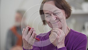Close-up face of smiling senior Caucasian woman looking at hand mirror. Female retiree looking back at blurred man and