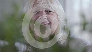 Close-up face of smiling senior Caucasian woman spraying water on domestic flowers. Portrait of happy retiree behind