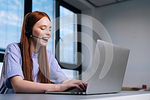 Close-up face of smiling young woman operator using headset and laptop computer during customer support.