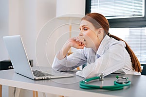 Close-up face of tired exhausted young female doctor wearing white coat yawning during working on laptop.