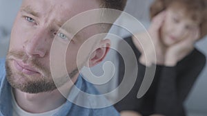 Close up face unhappy young man looking away in the foreground. The blurred figure of mature lady holding her head