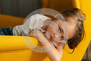 Close-up face of upset little girl with wet eyes from tears lying on yellow chair and looking at camera.