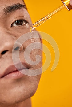 Close up of face of young asian man applying gel serum on his face using glass pipette isolated over yellow background