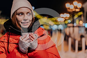 Close-up face of young woman wearing warm winter clothes enjoying drinking takeaway cup of coffee outdoors on cold