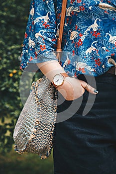 Close up, fashion blogger wearing a summer shirt and a white and brown analog wrist watch, holding a beautiful round straw purse.
