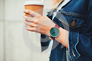 Close up fashion details, young fashionable woman in jeans jacket holding coffee cup. wearing casual unusual watch. Female in