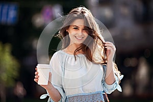 Close up fashion street style portrait of a beautiful girl in a casual outfit walks in the city . Beautiful brunette Keeps coffee.