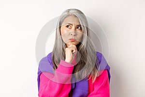 Close up of fashionable asian mature woman with grey hair, thinking and looking serious, looking aside while makign