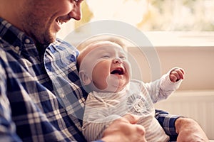 Close Up Of Father Cuddling Laughing Baby Son Sitting On Lap In Lounge Chair At Home Together