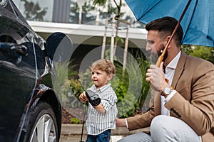 Close up of father and his little son charging their electric car on a rainy day, standing under umbrella. photo