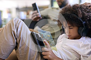 Close Up Of Father With Mobile Phone And Son Playing A Game On Digital Tablet With Headphones At Home