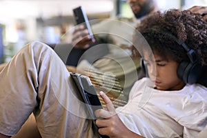 Close Up Of Father With Mobile Phone And Son Using Digital Tablet Reading With Headphones At Home