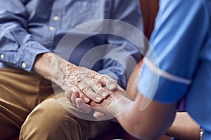 Close Up Of Female Care Worker In Uniform Holding Hands Of Senior Man Sitting In Care Home Lounge