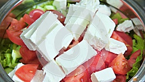 Close up of female chef hand putting Feta cheese cubes on greek vegan tomato salad with olives and lettuce leaves. The