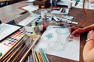 Close-up of female designer drawing floral compositions with crayons sitting at workplace surrounded with paint, gouache
